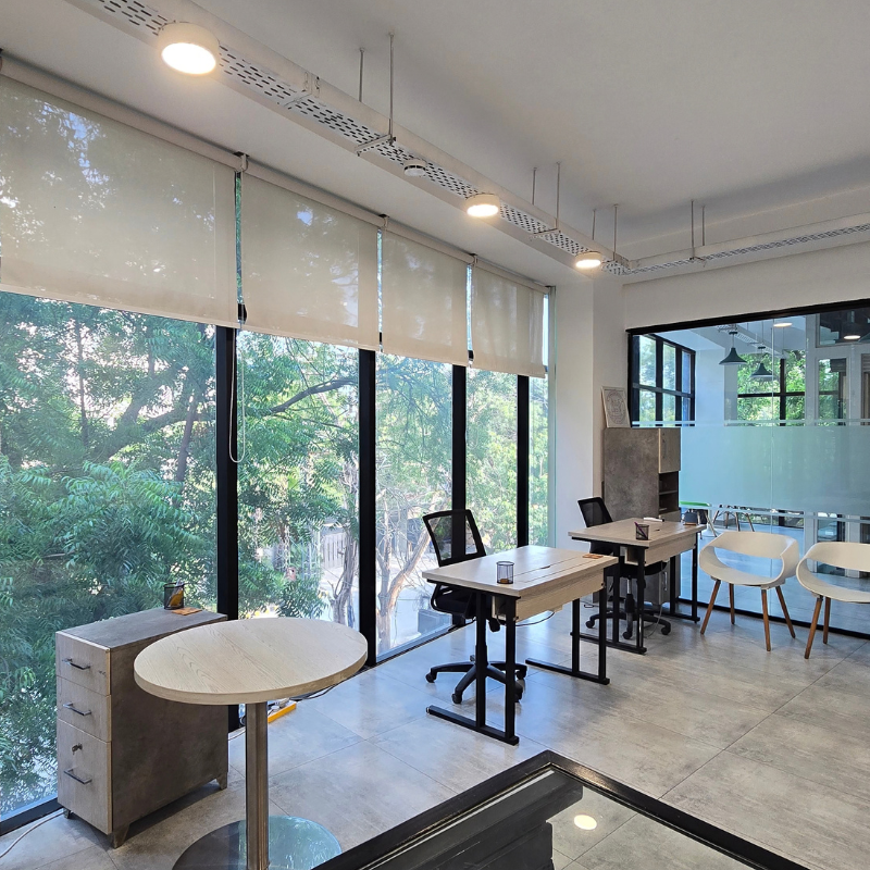 Coworking area with floor-to-ceiling windows, leafy trees outside, small wooden desks, and white chairs